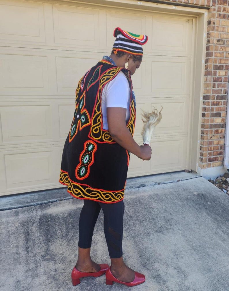 May include: A person wearing a black vest with colorful embroidery, a matching hat, and red shoes. The vest features intricate patterns in red, yellow, and green on a black background. The hat has a striped design with a curved top. The person is holding a white object.