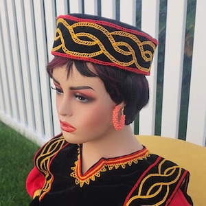 Pu&ograve; includere: Un cappello nero con un motivo ricamato rosso e giallo. Il cappello &egrave; indossato da un manichino, insieme a un capo nero e rosso con ricami gialli e orecchini a cerchio color corallo. Lo sfondo comprende una recinzione bianca ed erba verde.