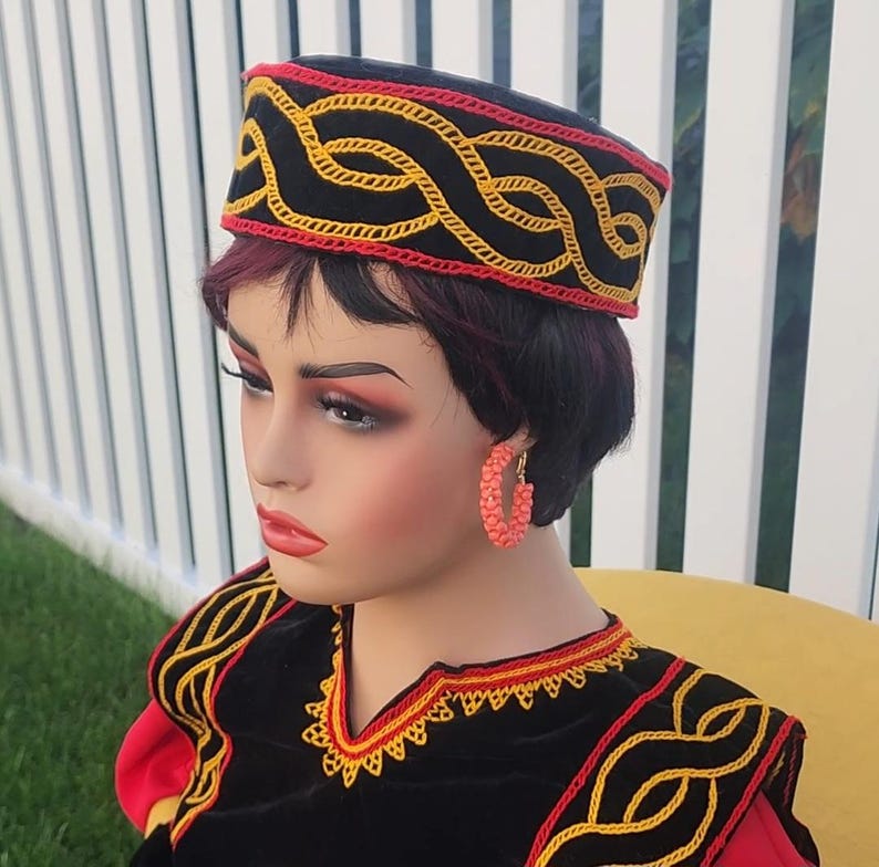 Pu&ograve; includere: Un cappello nero con un motivo ricamato rosso e giallo. Il cappello &egrave; indossato da un manichino, insieme a un capo nero e rosso con ricami gialli e orecchini a cerchio color corallo. Lo sfondo comprende una recinzione bianca ed erba verde.