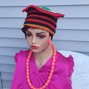 May include: A mannequin wearing a vibrant ensemble. The outfit includes a red, black, and yellow striped crochet hat, a pink beaded necklace, and matching earrings. The mannequin is also wearing a bright pink blouse.