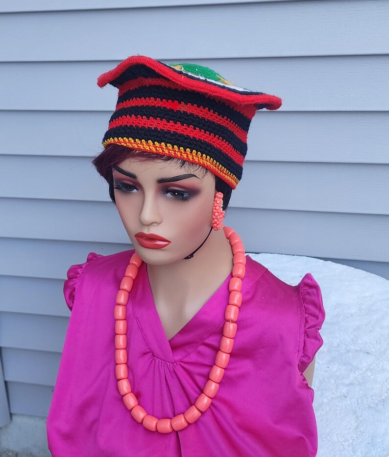 May include: A mannequin wearing a vibrant ensemble. The outfit includes a red, black, and yellow striped crochet hat, a pink beaded necklace, and matching earrings. The mannequin is also wearing a bright pink blouse.