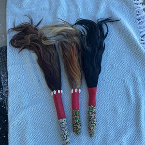 May include: Three decorative wands with hair-like strands in shades of brown, blonde, and black. Each wand has a red handle and is adorned with beads and shells. The wands are arranged on a light blue and white patterned fabric.