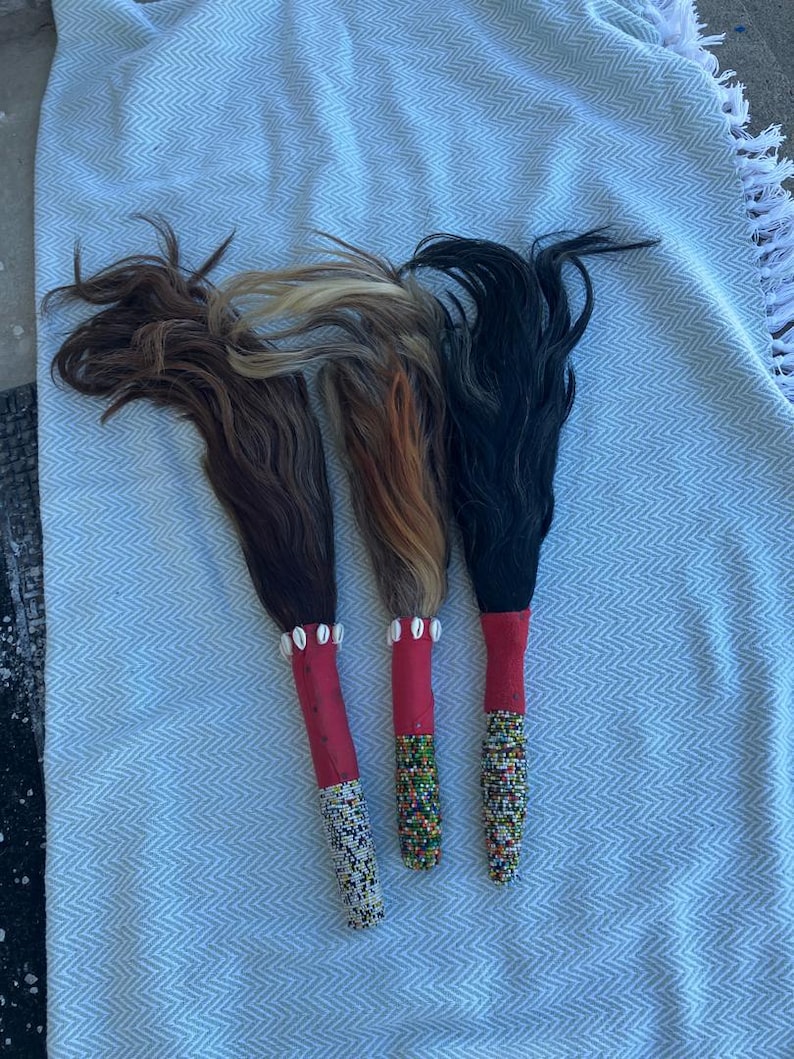 May include: Three decorative wands with hair-like strands in shades of brown, blonde, and black. Each wand has a red handle and is adorned with beads and shells. The wands are arranged on a light blue and white patterned fabric.