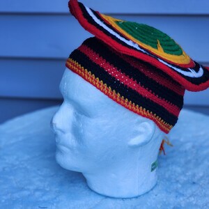May include: A colorful crochet hat with a wide brim, featuring red, black, yellow, green, and white stripes and accents. The hat is displayed on a white mannequin head, showcasing its unique design and craftsmanship.