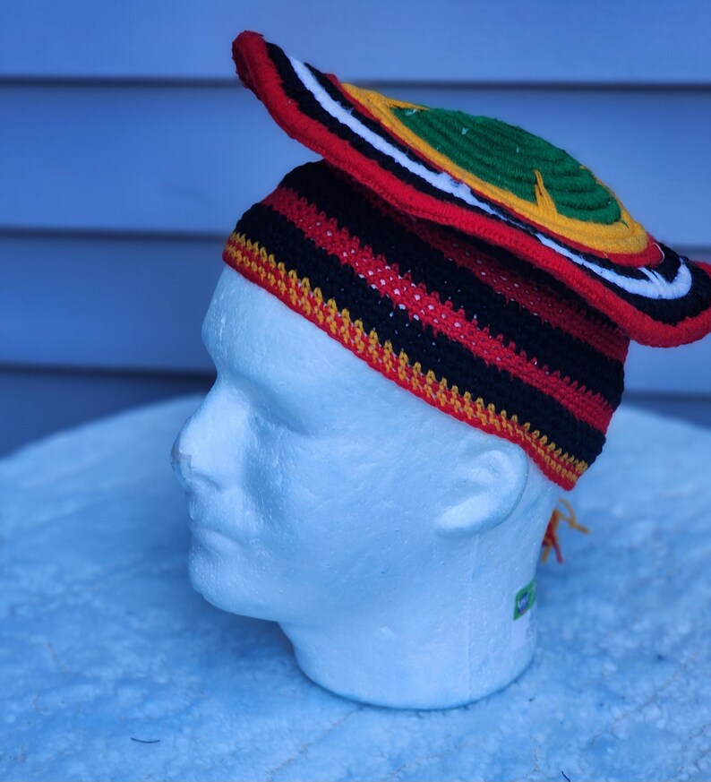 May include: A colorful crochet hat with a wide brim, featuring red, black, yellow, green, and white stripes and accents. The hat is displayed on a white mannequin head, showcasing its unique design and craftsmanship.