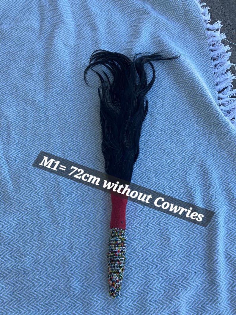 May include: A decorative item with a black, flowing top and a beaded handle. The handle is red and adorned with colorful beads. The text on the image reads "M1= 72cm without Cowries".