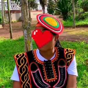 May include: A colorful, traditional hat with red, yellow, green, and white accents. The hat is worn with a black and red embroidered vest over a white shirt. The vest features intricate patterns and designs, showcasing cultural artistry.