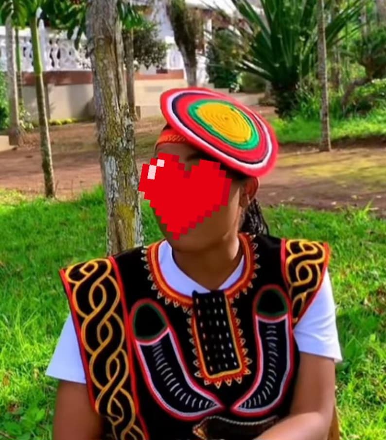 May include: A colorful, traditional hat with red, yellow, green, and white accents. The hat is worn with a black and red embroidered vest over a white shirt. The vest features intricate patterns and designs, showcasing cultural artistry.