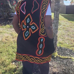 May include: A black vest with colorful embroidered patterns, including floral and geometric designs, is worn over a white shirt. The vest has a red, yellow, and black trim. A matching hat with a colorful design is also visible.
