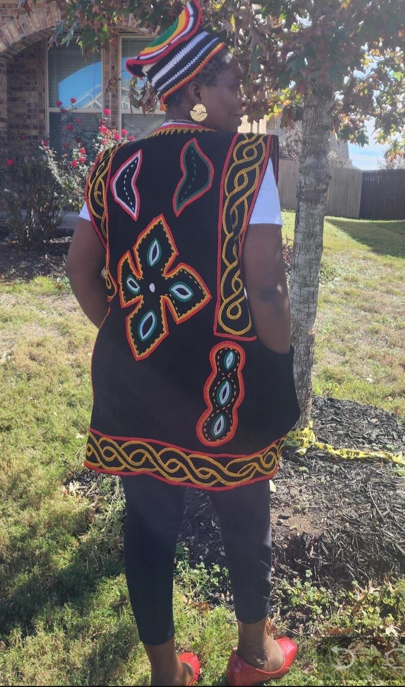 May include: A black vest with colorful embroidered patterns, including floral and geometric designs, is worn over a white shirt. The vest has a red, yellow, and black trim. A matching hat with a colorful design is also visible.