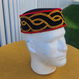 Pu&ograve; includere: Un cappello nero con un design ricamato rosso e oro. Il cappello ha una fascia di ricamo rosso e oro attorno alla base. Il cappello &egrave; esposto su una testa di manichino bianca.