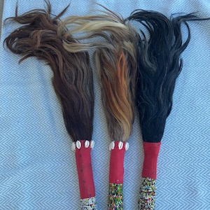 May include: Three decorative fly whisks with natural hair in shades of brown, blonde, and black. Each whisk features a red handle, adorned with white shells and colorful beaded patterns. The whisks are displayed on a light blue patterned surface.