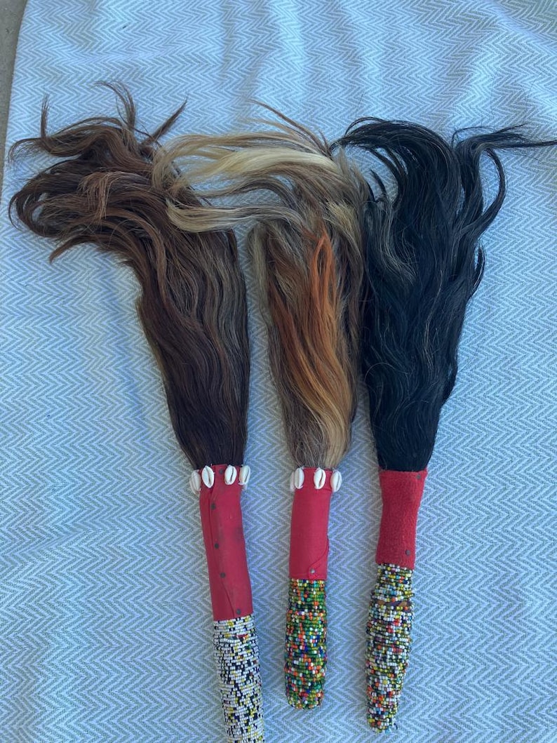 May include: Three decorative fly whisks with natural hair in shades of brown, blonde, and black. Each whisk features a red handle, adorned with white shells and colorful beaded patterns. The whisks are displayed on a light blue patterned surface.