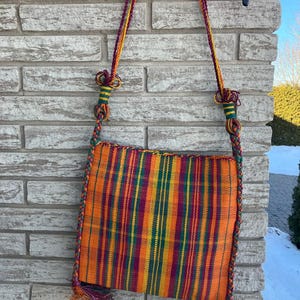 May include: A woven orange, green, yellow, and purple plaid crossbody bag with a braided strap and tassels.