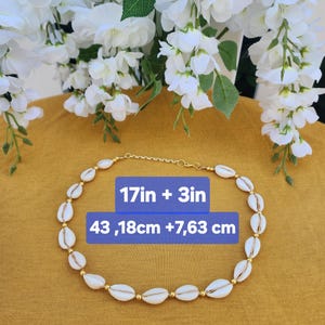 Może przedstawiać: Naszyjnik z białych muszli i złotych akcent&oacute;w. Naszyjnik ma około 43,18 cm plus 7,63 cm długości. Naszyjnik jest prezentowany na powierzchni w kolorze musztardowym, z białymi kwiatami w tle.