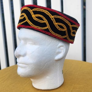 Pu&ograve; includere: Un cappello nero con un design ricamato rosso e giallo. Il cappello presenta un motivo ripetuto di forme gialle ad incastro, delineate da cuciture rosse. Il cappello &egrave; esposto su una testa di schiuma bianca.