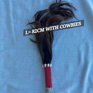 May include: A decorative item with a handle wrapped in red fabric and adorned with beads. The top features dark brown feathers and cowrie shells. The text on the image reads: L=82cm WITH COWRIES.