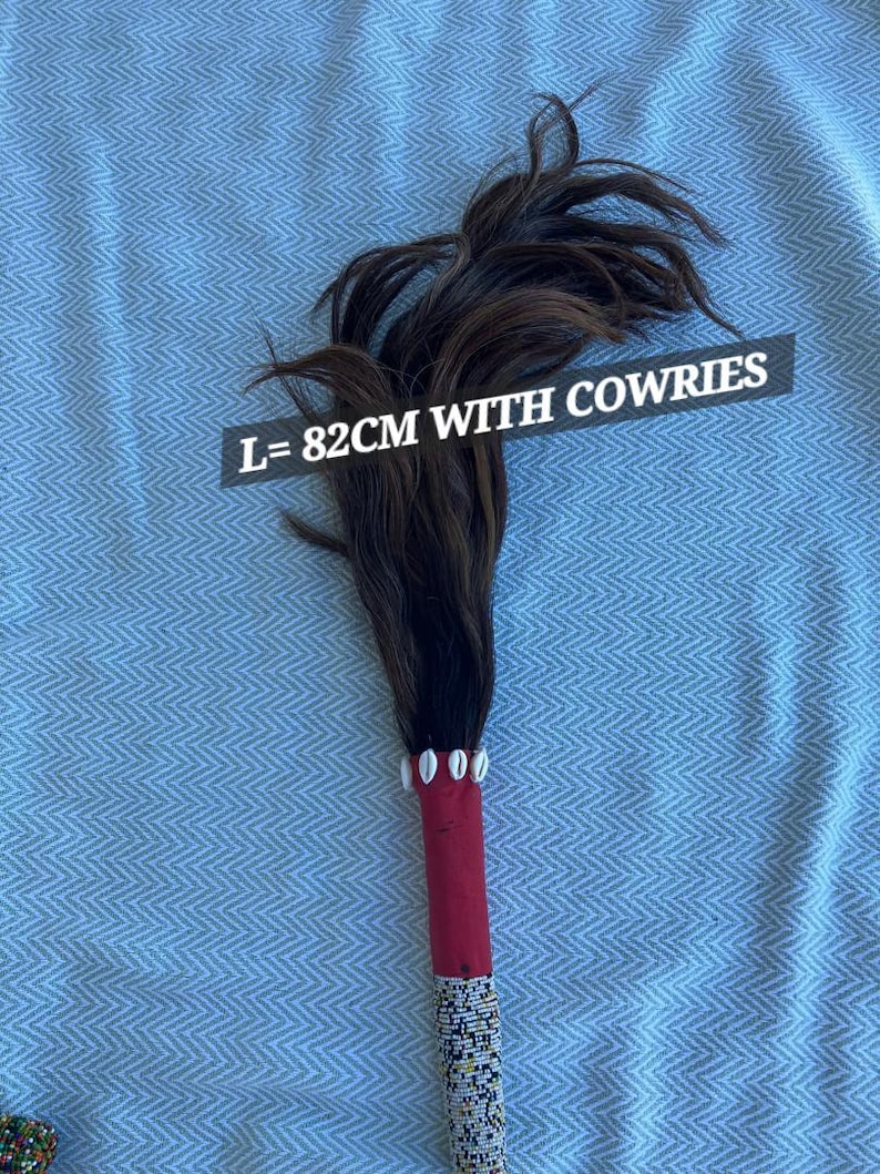 May include: A decorative item with a handle wrapped in red fabric and adorned with beads. The top features dark brown feathers and cowrie shells. The text on the image reads: L=82cm WITH COWRIES.