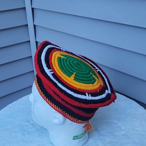May include: A colorful, handmade hat with a flat top. The hat features concentric circles of red, black, white, yellow, and green. The brim is red and black, and the hat has a small tassel. The hat is displayed on a white mannequin head.
