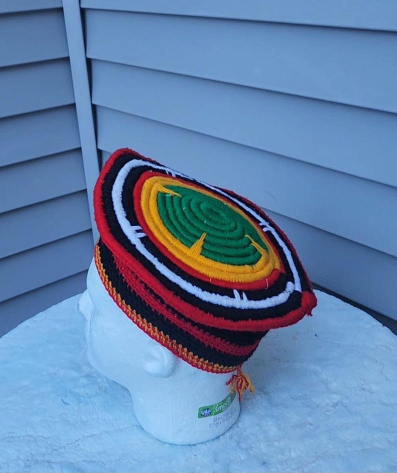 May include: A colorful, handmade hat with a flat top. The hat features concentric circles of red, black, white, yellow, and green. The brim is red and black, and the hat has a small tassel. The hat is displayed on a white mannequin head.