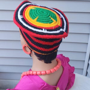 May include: A colorful, handmade crochet hat with concentric circles of red, black, white, yellow, and green. The hat is worn with matching coral-colored jewelry and a bright pink dress. The hat's design is eye-catching and vibrant.