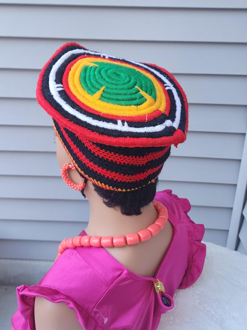 May include: A colorful, handmade crochet hat with concentric circles of red, black, white, yellow, and green. The hat is worn with matching coral-colored jewelry and a bright pink dress. The hat's design is eye-catching and vibrant.
