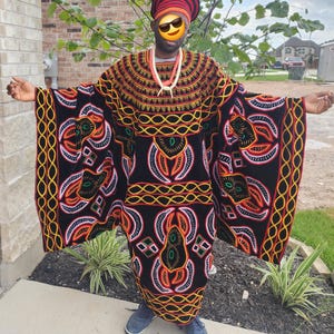 May include: A person wearing a traditional African outfit. The black garment is adorned with intricate patterns in orange, yellow, and white. A matching head covering and beaded necklace complete the ensemble. The person is also wearing blue shoes.