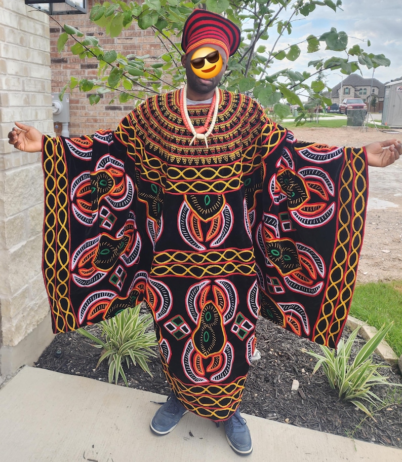 May include: A person wearing a traditional African outfit. The black garment is adorned with intricate patterns in orange, yellow, and white. A matching head covering and beaded necklace complete the ensemble. The person is also wearing blue shoes.