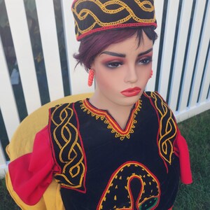 Pu&ograve; includere: Un manichino indossa un completo ricamato nero e rosso, compreso un cappello e un top. Il cappello &egrave; nero con ricami gialli e rossi. Il top &egrave; nero con ricami gialli e maniche rosse. Il manichino indossa anche orecchini rossi.