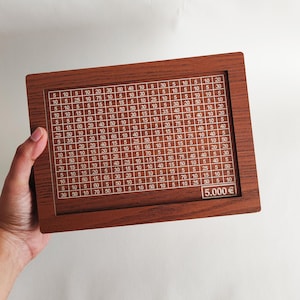 Puede incluir: Un tablero de madera con una cuadrícula de números, cada casilla contiene una cantidad de dinero diferente. El tablero es marrón con un borde blanco y los números son blancos. El tablero tiene una etiqueta en la parte inferior que dice "5.000 €".