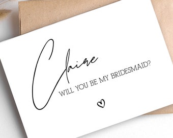 May include: A white card with black text that reads "Claire, Will you be my bridesmaid?" with a small heart symbol below the text.