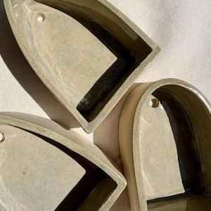 May include: Three handmade, beige ceramic soap dishes. Each dish has a unique shape with a curved top, a flat bottom, and a small hole. The interior of each dish has a dark, contrasting color.