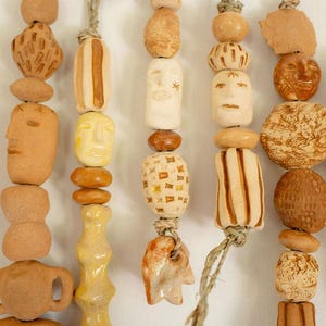 May include: A collection of vertical hanging strands featuring various hand-sculpted clay beads in shades of tan, beige, and brown. The beads display diverse textures, geometric patterns, and carved human face motifs, all strung together on natural fiber twine.