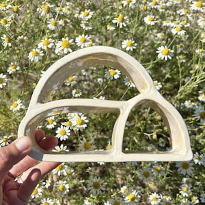 Pode incluir: Uma escultura de cerâmica na cor creme com um design de moldura aberta arqueada, uma divisória horizontal e uma seção arqueada menor no lado direito. O objeto tem um acabamento liso e brilhante e uma forma estrutural oca com bordas curvas.
