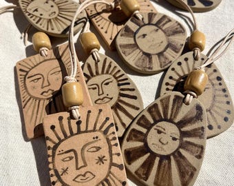 May include: A set of eight ceramic sun-shaped ornaments with hand-painted faces and brown accents. Each ornament has a brown leather cord with a wooden bead.
