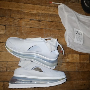 May include: White sneakers with a distinctive design, including a strap and cut-out detail. The soles are transparent, showcasing an air cushion. A white bag with the number "720" is also visible.