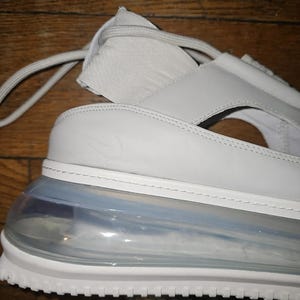 May include: A close-up of a light grey sandal with a white sole and a clear, air-cushioned heel. The sandal features a strap design and the Nike swoosh logo. The shoe is made of leather and synthetic materials.