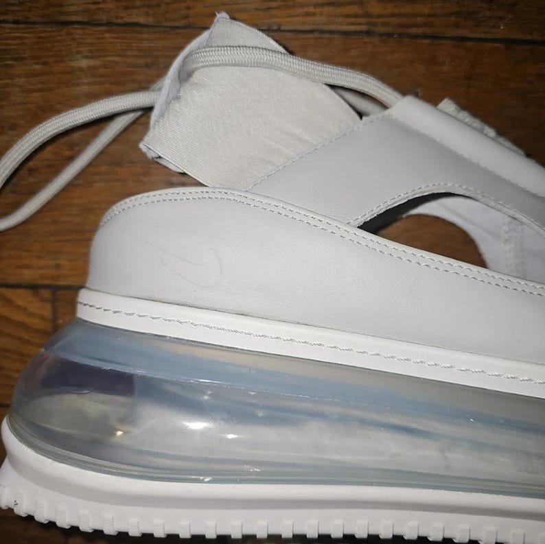 May include: A close-up of a light grey sandal with a white sole and a clear, air-cushioned heel. The sandal features a strap design and the Nike swoosh logo. The shoe is made of leather and synthetic materials.