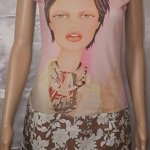 Peut inclure: Ensemble deux pièces composé d'un haut sans manches et d'une jupe. Le haut présente un portrait de femme aux cheveux foncés sur fond rose. La jupe est marron avec un motif floral blanc et bleu clair. Le haut a une bordure bleu clair.