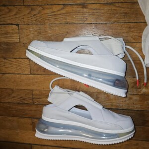 May include: Two light grey sneakers with a distinctive design. The shoes feature a clear air bubble sole, white soles, and a strap across the top. The laces have red tips. The shoes are on a wooden surface.