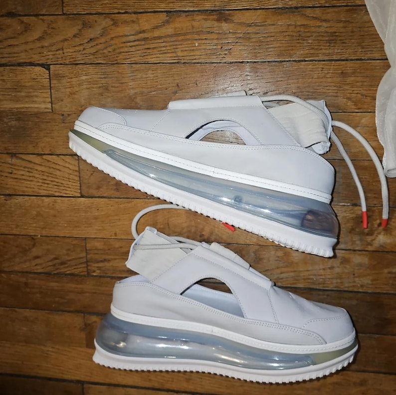 May include: Two light grey sneakers with a distinctive design. The shoes feature a clear air bubble sole, white soles, and a strap across the top. The laces have red tips. The shoes are on a wooden surface.