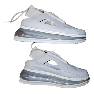 May include: Two pairs of white trainers with a distinctive design. The shoes feature a light grey upper with a cut-out and a clear, bubble-like sole. The laces are thin and white. The shoes are set against a white background.