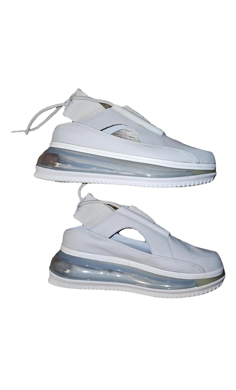 May include: Two pairs of white trainers with a distinctive design. The shoes feature a light grey upper with a cut-out and a clear, bubble-like sole. The laces are thin and white. The shoes are set against a white background.