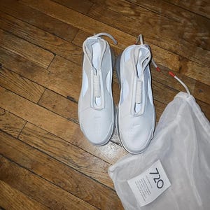 May include: Light grey sneakers with a distinctive design, featuring a zip closure and a minimalist aesthetic. The shoes are on a wooden surface, with a white drawstring bag and a tag with the number "720".