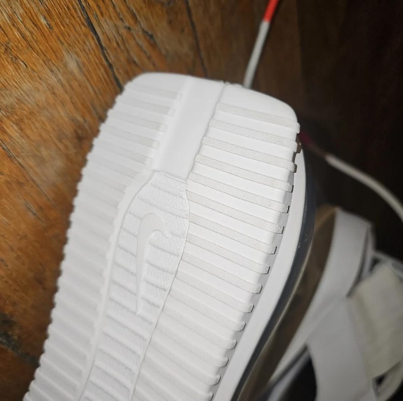 May include: Close-up of the white sole of a sandal. The sole has a textured, ridged pattern with the Nike logo. The sandal's design includes a thick sole and a strap, suggesting a sporty or casual style.