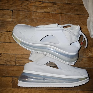 May include: Pair of white sandals with a unique design. The sandals feature a thick, transparent sole with a visible air pocket, and a white leather upper with a strap across the top. The soles have a silver trim.