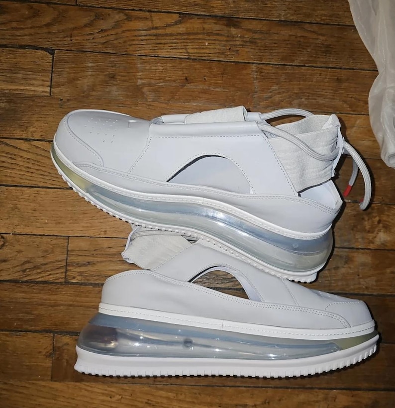 May include: Pair of white sandals with a unique design. The sandals feature a thick, transparent sole with a visible air pocket, and a white leather upper with a strap across the top. The soles have a silver trim.
