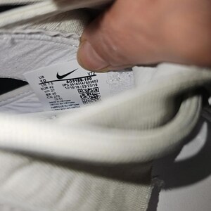 May include: A close-up of a white trainer's interior, displaying a label with the Nike logo and product details. The label includes size information: US 10, UK 7.5, EUR 42, and CM 27. The style code is A03189-100.