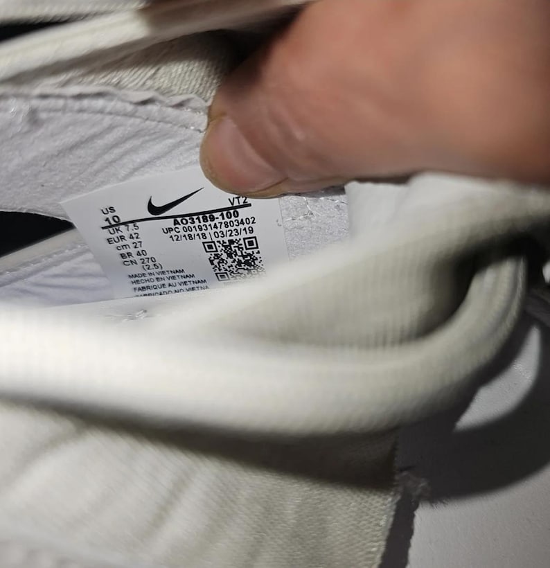 May include: A close-up of a white trainer's interior, displaying a label with the Nike logo and product details. The label includes size information: US 10, UK 7.5, EUR 42, and CM 27. The style code is A03189-100.