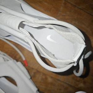 May include: Close-up of a white trainer with a visible Nike logo on the insole. The shoe features white laces and a textured fabric strap. The design includes cut-out sections and a rounded toe.